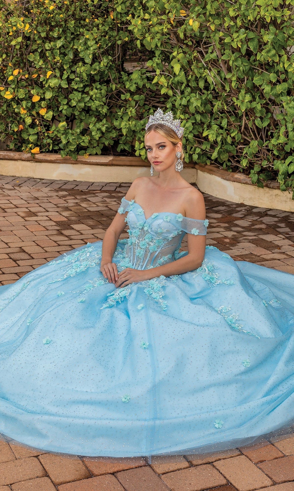 A woman in the Quinceanera Dress 1867 By Dancing Queen, an aqua blue off-the-shoulder ball gown, sits outdoors on a brick surface with lush green bushes behind her, wearing a sparkling tiara.